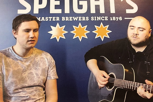 Photo of two men, one with guitar, in front of Speights sign