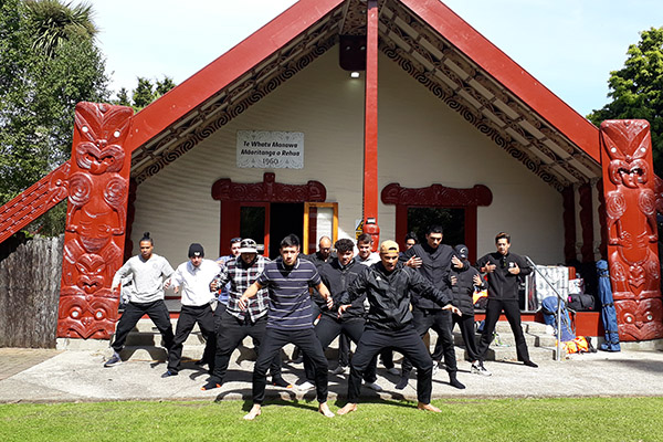 Photo of a group of young men doing a haka outside a marae.