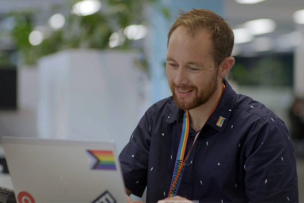 Man in rainbow lanyard working at laptop with trans pride sticker on
