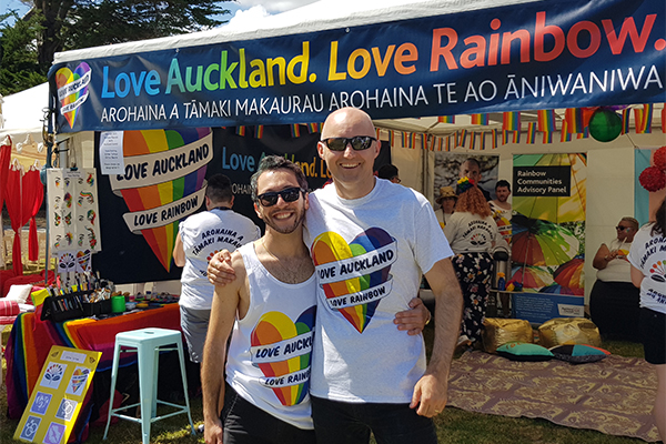 Two men hugging in front of rainbow tent at Auckland Pride