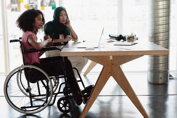Two women, one in a wheelchair, the other wearing a heaopdscarf, looking at resources together on a lapt